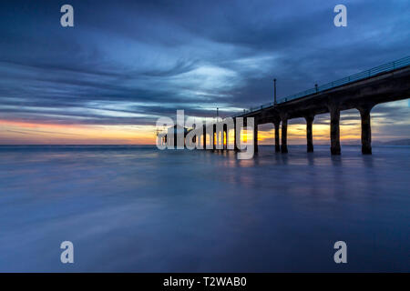Long-exposure shot of colorful sky and clouds over Manhattan Beach Pier at sunset with smooth waves washing onto the beach, Manhattan Beach, Californi Stock Photo
