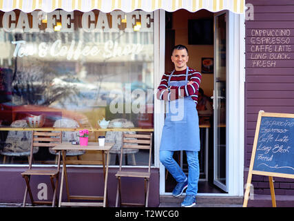 Portrait of a confident young man standing in the doorway of a coffee shop. Tea and Coffee Shop written on the window. Traffic reflection. His welcomi Stock Photo