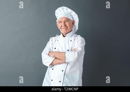 Senior male chef studio standing isolated on gray wall crossed arms confident looking camera smiling cheerful Stock Photo