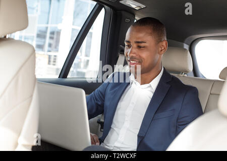 Young african american businessman sitting on the back seat in the car browsing internet on laptop smiling joyful Stock Photo