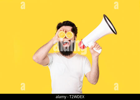Happy bearded hipster man with sliced lemon on eyes, shouting through a megaphone, over yellow background, concept of summer mood Stock Photo