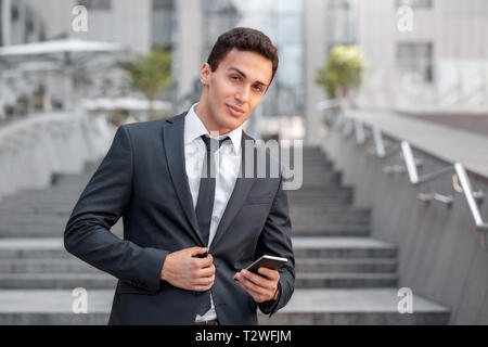 Young business man standing on the city street holding smartphone using application looking camera smiling playful Stock Photo