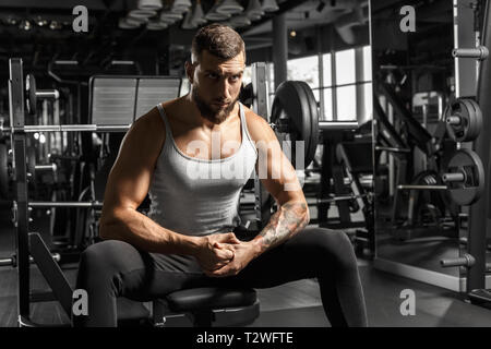 Bearded man bodybuilder sitting on bench at gym looking forward motivated Stock Photo