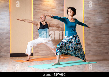 Man and woman with outstretched arms are practicing yoga while standing on blue mat in gym. Stock Photo
