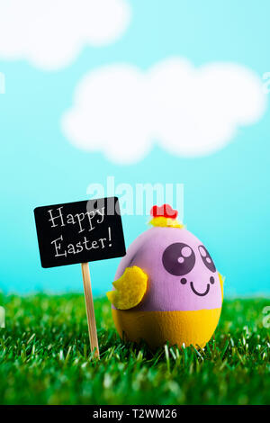 a handmade easter egg, decorated as a chick with a cute and funny face, on the grass, next to a black signboard with the text happy easter written in  Stock Photo