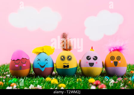 closeup of some handmade easter eggs, with cute and funny faces and hairstyles, on the grass with a pink sky with white clouds in the background Stock Photo