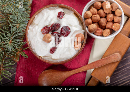 Wooden and ceramic bowls of yogurt with dried cranberries and hazelnuts, spoon and fir twig on red kitchen napkin on tray on wooden table. Top view. Stock Photo