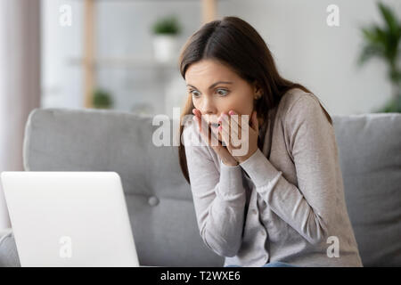 Woman sitting on couch reading message on computer feels shocked Stock Photo