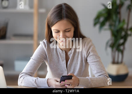 Joyful woman sitting at desk reading message on phone Stock Photo