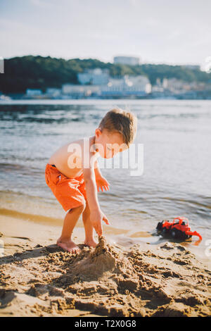 Subject construction and heavy industry. Abstraction child boy playing on the sand near the river in the summer toy red tractor model, excavator machi Stock Photo