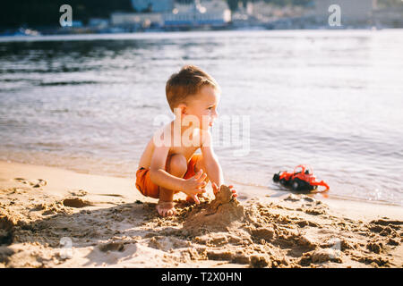 Subject construction and heavy industry. Abstraction child boy playing on the sand near the river in the summer toy red tractor model, excavator machi Stock Photo