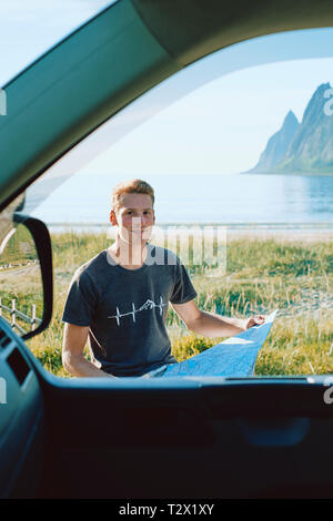 A location portrait at the coast of a young millennial living and traveling in his camper van reading a map and planning his next route - on the road Stock Photo