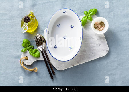 empty ceramic white baking dish on a marble cutting board and cutlery on a blue linen tablecloth with olive oil, spices and basil leaves. home cooking Stock Photo