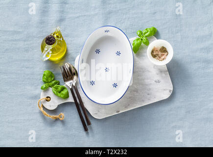 empty ceramic white baking dish on a marble cutting board and cutlery on a blue linen tablecloth with olive oil, spices and basil leaves. home cooking Stock Photo