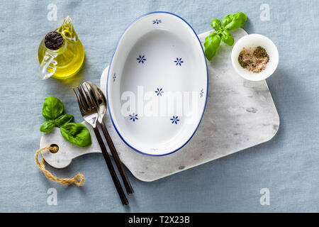 empty ceramic white baking dish on a marble cutting board and cutlery on a blue linen tablecloth with olive oil, spices and basil leaves. home cooking Stock Photo