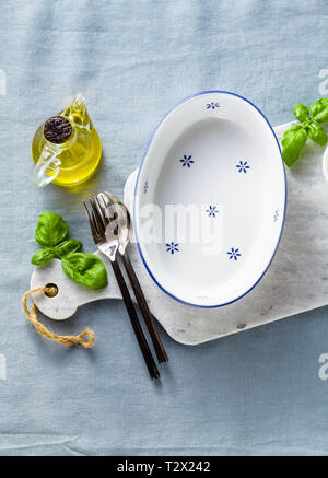 empty ceramic white baking dish on a marble cutting board and cutlery on a blue linen tablecloth with olive oil, spices and basil leaves. home cooking Stock Photo
