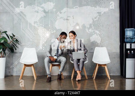 multiethnic colleagues with laptop and coffee to go sitting and checking time on watch in waiting hall with map illustration on wall Stock Photo