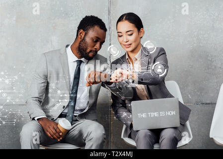 multiethnic colleagues with laptop and coffee sitting and looking at smartwatches in waiting hall with the time is now lettering on laptop and notific Stock Photo