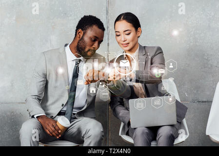 multiethnic colleagues with laptop and coffee sitting and looking at smartwatches in waiting hall with notifications icons Stock Photo
