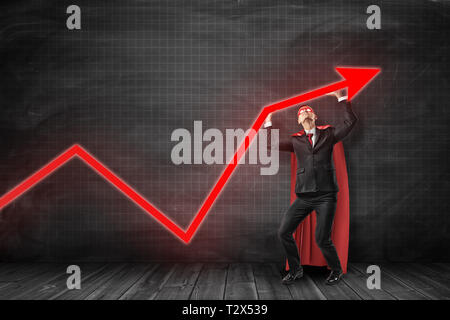 Businessman in black suit and red cape and red eye mask looking at and holding up red graph arrow pointing up at black graph ruled wall. Business supe Stock Photo