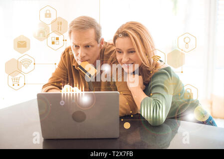 concentrated man holding credit card with lips while using laptop together with smiling wife, smart home concept Stock Photo