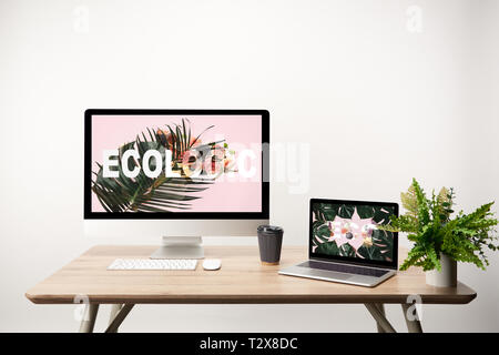 computer with ecologic illustration on monitor and laptop with monstera leaves and eco lettering on screen on wooden table Stock Photo