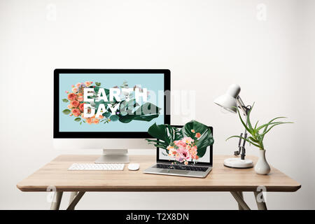 computer with earth day illustration on monitor and laptop with monstera leaves and flowers illustration on screen on wooden table Stock Photo