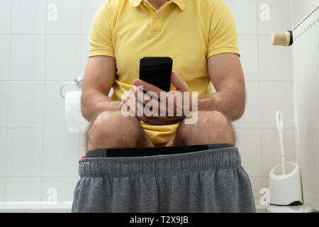 Mid-section Of Man Sitting On Toilet Using Smartphone Stock Photo