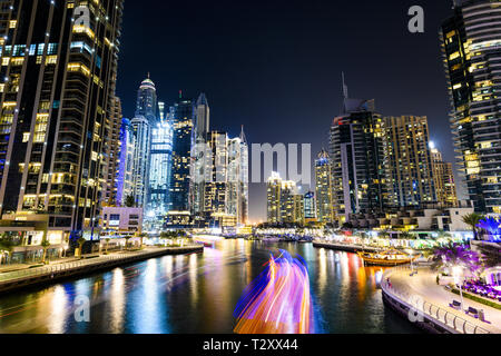 Stunning view of the Dubai Marina at dusk with illuminated skyscrapers in the background and a light trails left by a yacht sailing in the foreground. Stock Photo