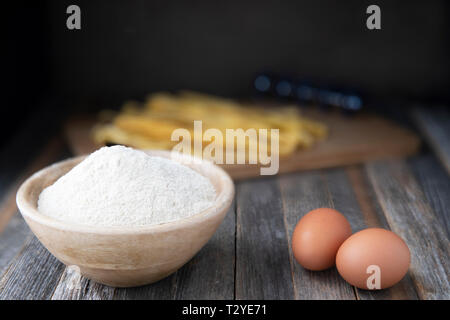 Bowl of whole wheat flour and two eggs on a rustic wooden table with pappardelle pasta in the background. Stock Photo