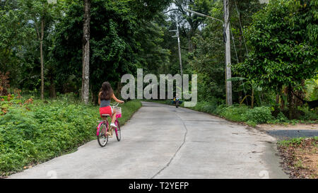 Girl in red shorts riding bicycle on road in village in rain forest Stock Photo