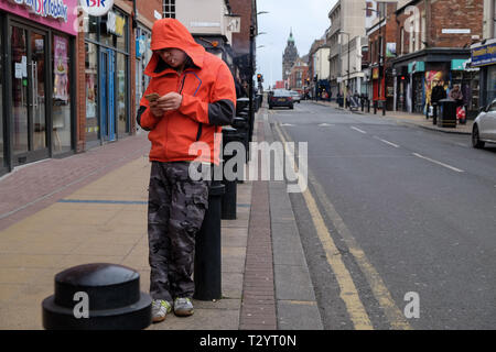 Asian man smoking a cigarette and texting on his phone while leaning on a bollard on a city street Stock Photo