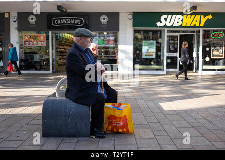 Older man in cloth cap. with walking stick and shopping bag, sitting on a concrete bench on the Moor pedestrian precinct in Sheffield Stock Photo