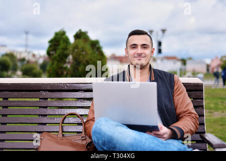portrait of young guy. Freelancer working on laptop in the street. happy smiling man sitting on bench Stock Photo