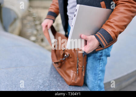young guy freelancer in jacket puts laptop in leather bag Stock Photo