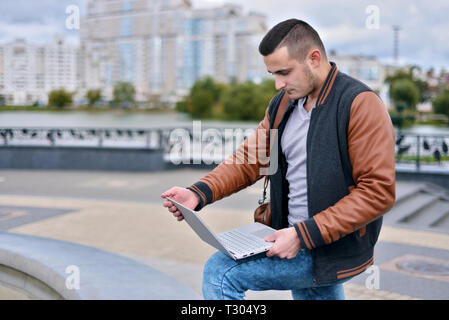 young guy successful freelancer in leather jacket working on the street. man holding laptop on his lap Stock Photo