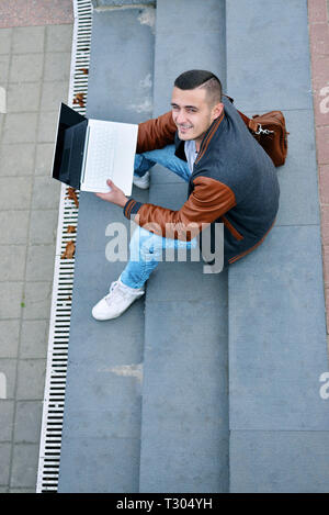 young freelance guy. happy, successful one in a leather jacket working on a laptop on the street. man sits on the stairs and looks up and smiles. view Stock Photo