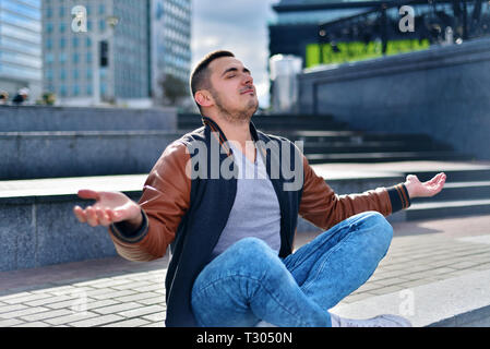 young freelance guy in leather jacket relaxes and meditates on the street. man sits on the stairs in the city in the Zen position Stock Photo