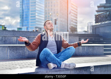 young freelance guy in leather jacket and sneakers, relaxes and meditates on the street. man sits on the stairs in big city in a zen position Stock Photo