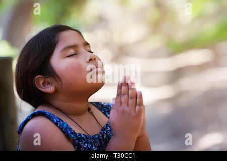 A young girl praying with hands together in reverence to God, wearing conservative clothing and in an outdoor setting. Stock Photo