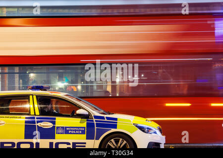London metropolitan police car waiting at a traffic light on a night patrol with double decker bus passing, in motion, on the background. Stock Photo