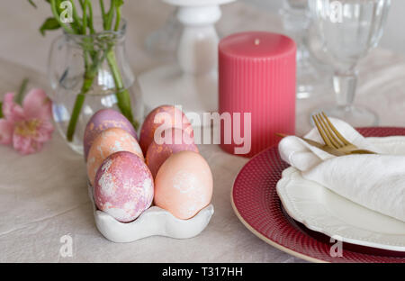 Close up Easter colorful eggs at concrete eggs holder on linen tablecloth with serving plate and candles. Concept of Ester food. Stock Photo