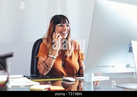 Smiling businesswoman talking on mobile phone while looking at a computer. Cheerful entrepreneur working in office sitting at her desk. Stock Photo