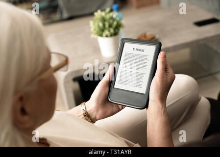 Senior woman sitting by the window, reading an e-book on digital tablet device Stock Photo