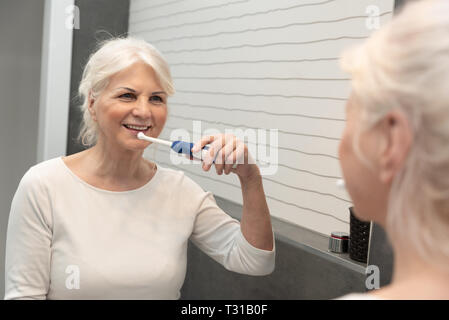 Cute mature woman brushing teeth. Electric toothbrush used by senior woman. Stock Photo