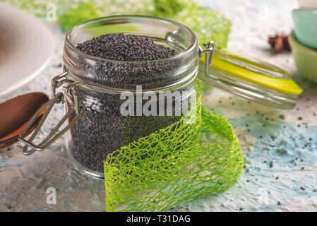 Horizontal photo of glass jar with sealed opened cover. Jar is full of raw poppy seeds. Jar is placed on light blue and white textured board. Green ri Stock Photo