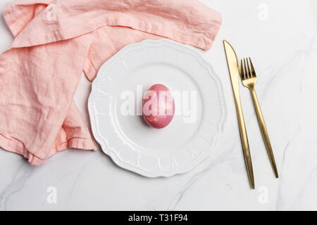 Easter table, place setting. Easter egg, pink color painted in a white plate, pink napkin, golden tableware on white marble background, horizontal, co Stock Photo