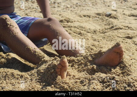 Boy in shorts with legs partly covered in sand at the beach Stock Photo
