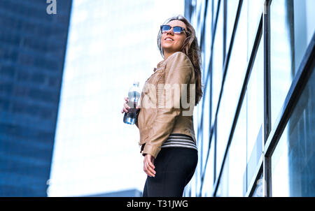 Confident woman in city with water bottle. Trendy lady standing next to modern glass business buildings wind in hair. Active healthy lifestyle. Stock Photo