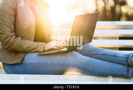 Woman using laptop in sunset outdoors. Lady writing with computer while sitting on park bench in sunset. Remote work or studying in nature concept. Stock Photo
